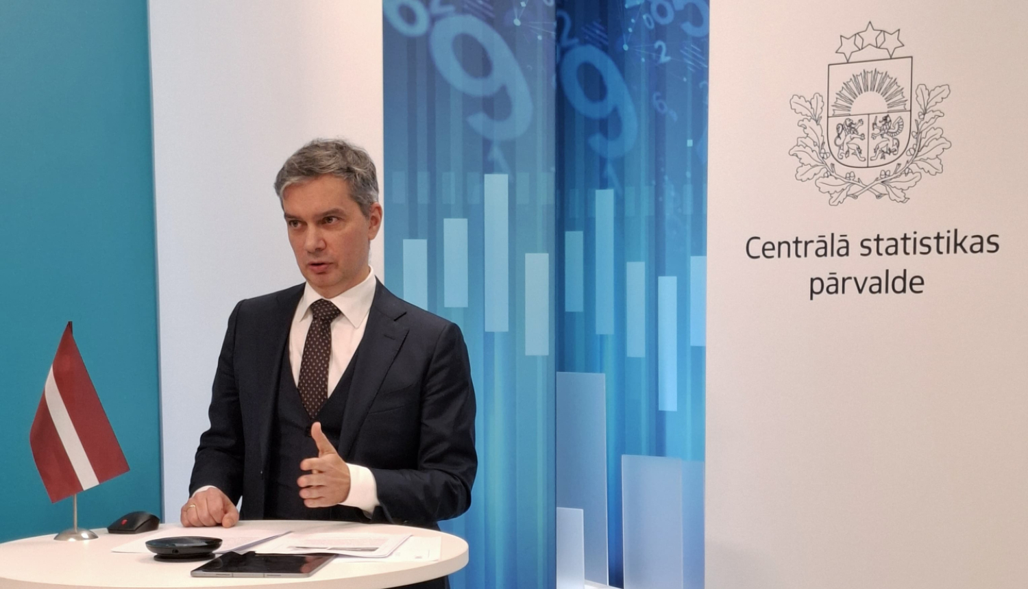 Raimonds Lapiņš the Director General of the CSB speaks at the table. On the table is a small Latvian flag, a tablet and documents. In the background to the right - the coat of arms and the full name of the CSB.