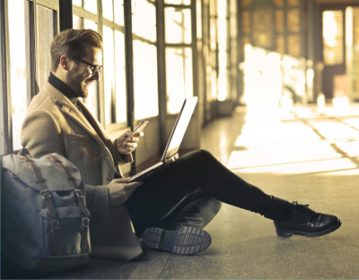 Vīrietis sēž ar klēpī datoru, rokās mobilo telefonu