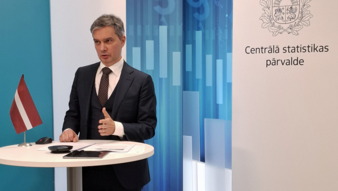 Raimonds Lapiņš the Director General of the CSB speaks at the table. On the table is a small Latvian flag, a tablet and documents. In the background to the right - the coat of arms and the full name of the CSB.