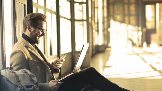 Vīrietis sēž ar klēpī datoru, rokās mobilo telefonu