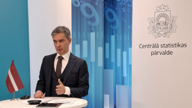 Raimonds Lapiņš the Director General of the CSB speaks at the table. On the table is a small Latvian flag, a tablet and documents. In the background to the right - the coat of arms and the full name of the CSB.