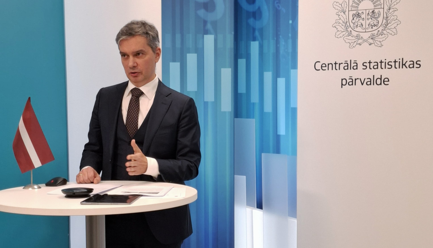 Raimonds Lapiņš the Director General of the CSB speaks at the table. On the table is a small Latvian flag, a tablet and documents. In the background to the right - the coat of arms and the full name of the CSB.